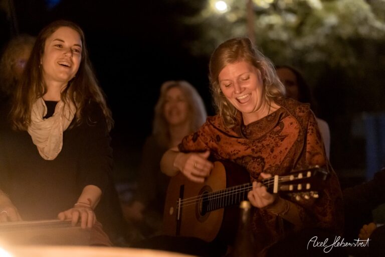 Zwei Frauen am Lagerfeuer, eine spielt Gitarre, die andere lacht, entspannte und fröhliche Atmosphäre in der Natur – © Axel Hebenstreit
