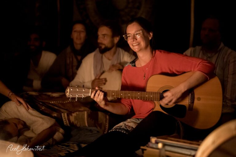 Eine lächelnde Frau spielt in einer gemütlichen, abgedunkelten Umgebung Gitarre, während andere aufmerksam lauschen – © Axel Hebenstreit