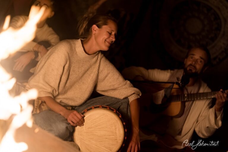 Frau spielt auf einer Trommel und lächelt am Lagerfeuer, während ein Mann Gitarre spielt in einer gemütlichen Atmosphäre – © Axel Hebenstreit