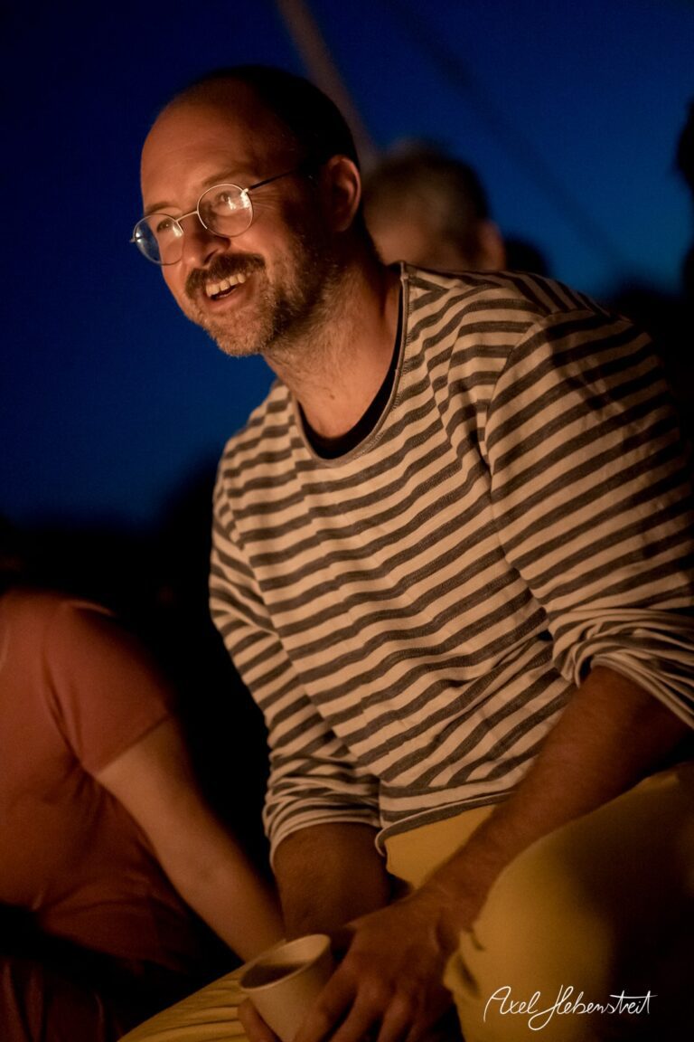 Ein lächelnder Mann in gestreiftem Oberteil sitzt am Lagerfeuer und genießt die gemütliche Atmosphäre – © Axel Hebenstreit