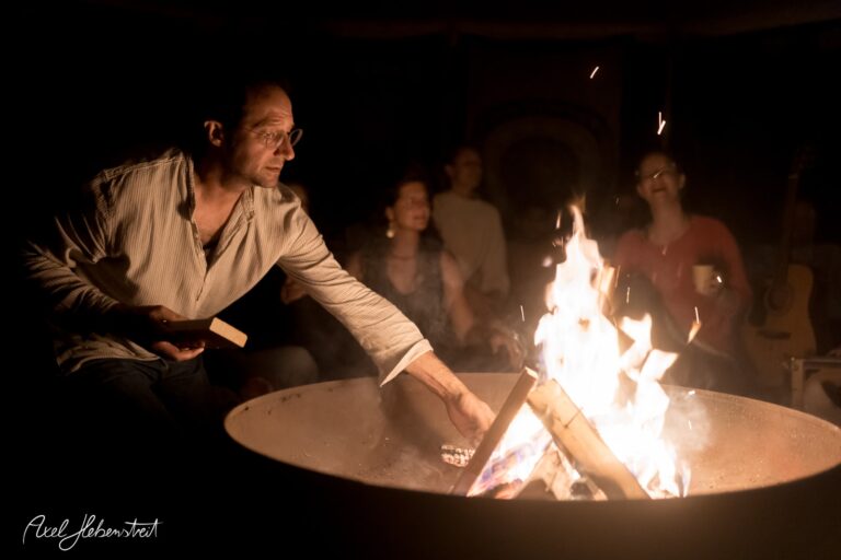 Ein Mann fügt Holz in ein Lagerfeuer hinzu, während eine Gruppe von Menschen in der warmen Atmosphäre zusammen sitzt – © Axel Hebenstreit
