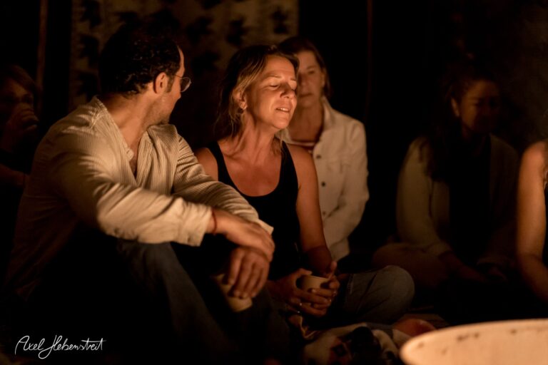 Eine Gruppe von Menschen sitzt in einem gedämpften Licht, vertieft in ein Gespräch und Genuss von Getränken. – © Axel Hebenstreit