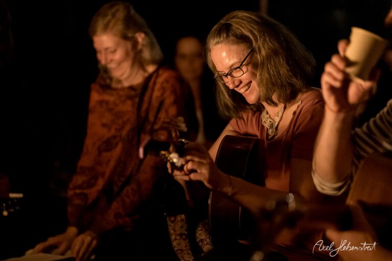 Zwei Frauen lachen und musizieren am Lagerfeuer, umgeben von einer warmen, festlichen Atmosphäre – © Axel Hebenstreit