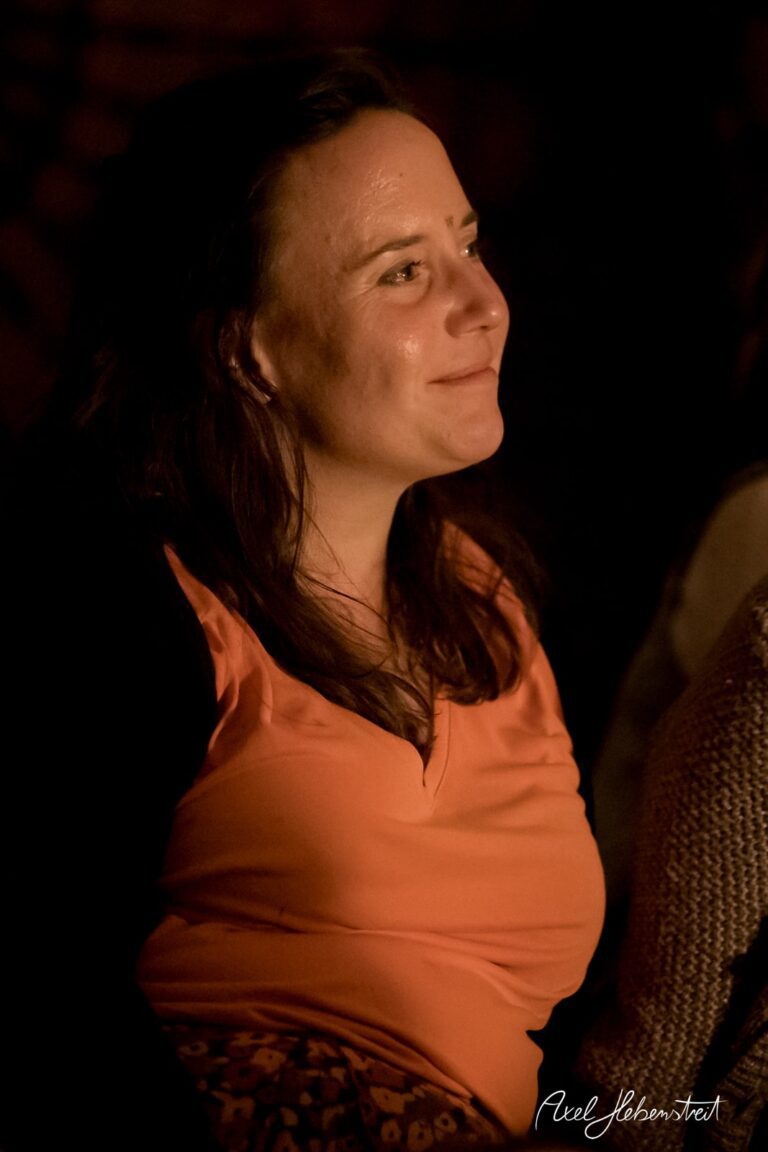 Frau in orangefarbener Bluse, lächelnd im warmen Licht, während sie eine Veranstaltung genießt – © Axel Hebenstreit