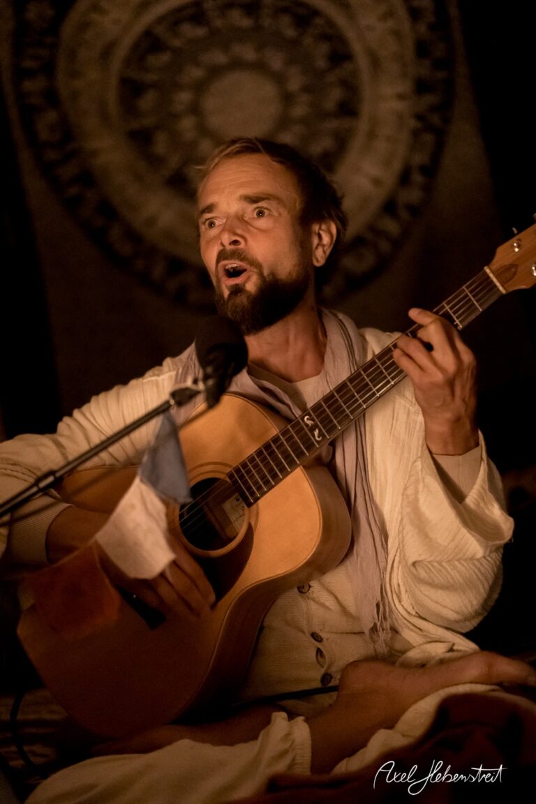 Ein Musiker mit Gitarre singt leidenschaftlich in einer warmen, intimen Atmosphäre – © Axel Hebenstreit