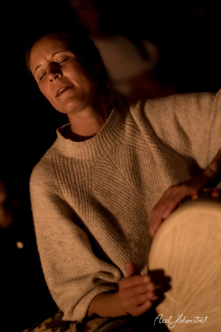 Eine Frau spielt mit geschlossenen Augen auf einer Trommel, umgeben von einer warmen, feierlichen Atmosphäre – © Axel Hebenstreit.