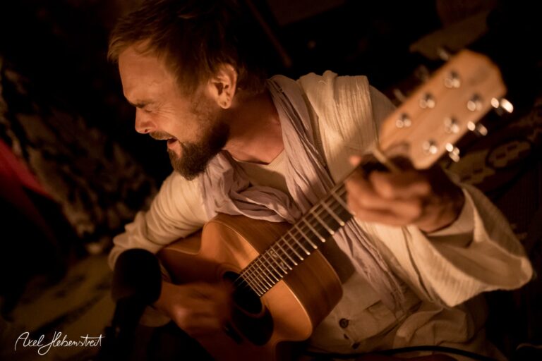 Ein Musiker singt leidenschaftlich mit einer Gitarre in einer stimmungsvollen, gedämpften Beleuchtung – © Axel Hebenstreit.