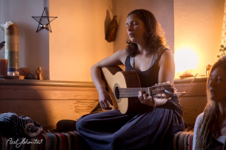 Frau mit Gitarre sitzt in einem stimmungsvollen Raum und singt, umgeben von sanftem Licht – © Axel Hebenstreit