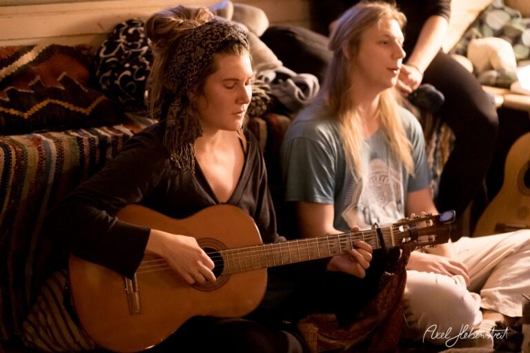 Eine Musikerin spielt Gitarre, während eine entspannte Atmosphäre im Hintergrund herrscht – © Axel Hebenstreit.