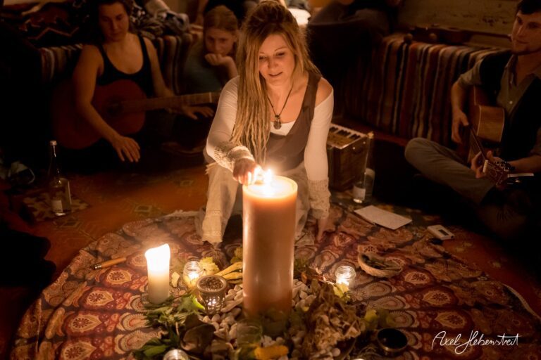 Eine Frau zündet eine große Kerze in einer geselligen Runde an, umgeben von Kerzenlicht und Naturmaterialien – © Axel Hebenstreit.