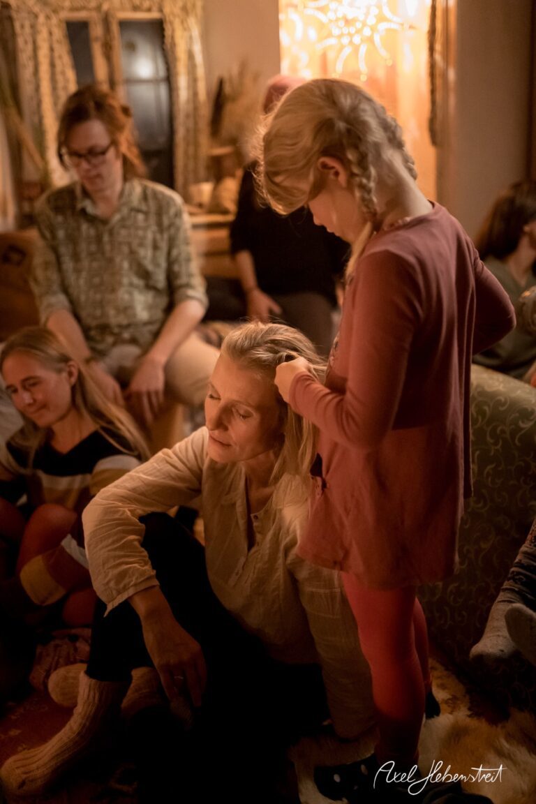 Ein Mädchen flechtet einer Frau im stimmungsvollen Wohnzimmer die Haare, während andere entspannt zuschauen – © Axel Hebenstreit
