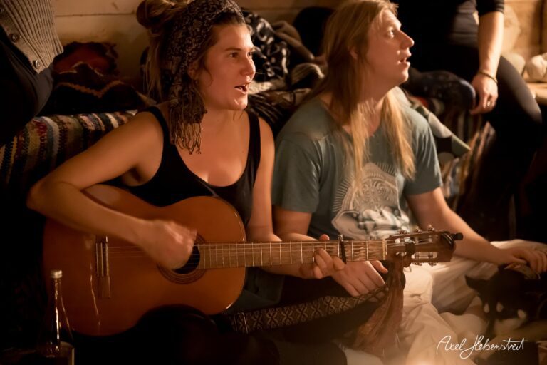 Musikabend in gemütlicher Runde: Eine Frau spielt Gitarre und singt, während ein Mann aufmerksam zuhört. – © Axel Hebenstreit