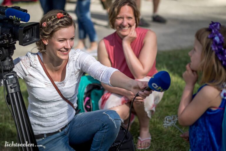 Eine junge Reporterin interviewt ein Mädchen während eines Festivals im Freien, umgeben von fröhlicher Stimmung.