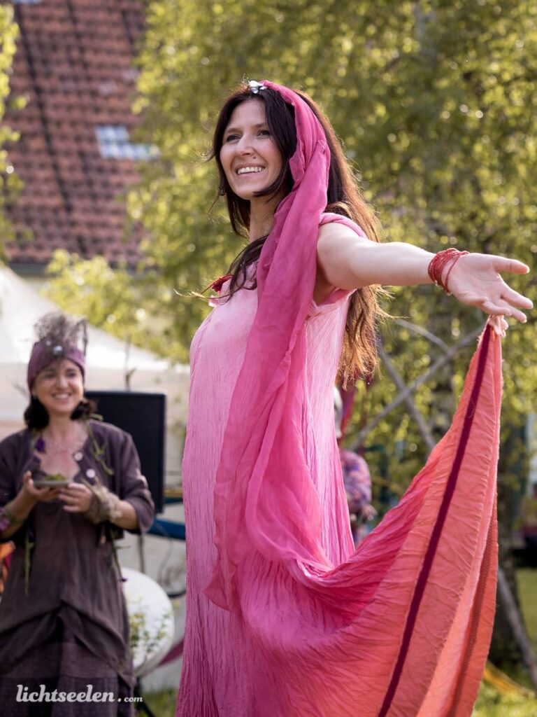 Eine Frau in einem pinken Kleid tanzt fröhlich auf einem Festival im Freien, umgeben von Bäumen und fröhlichen Menschen.
