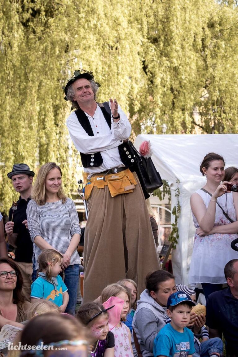 Ein großgewachsener, kostümierter Künstler steht auf einer Bank und applaudiert begeistert im Festivalkontext.