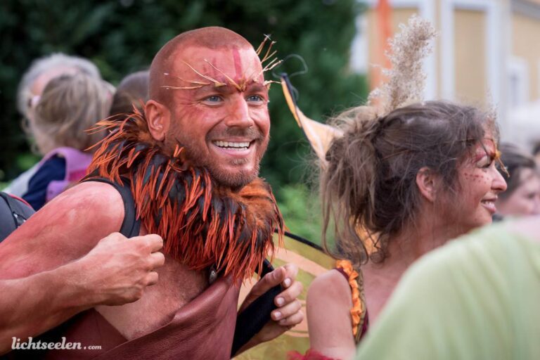 Mann mit auffälligem Kostüm und Körperbemalung lächelt fröhlich auf einem Festival im Freien.