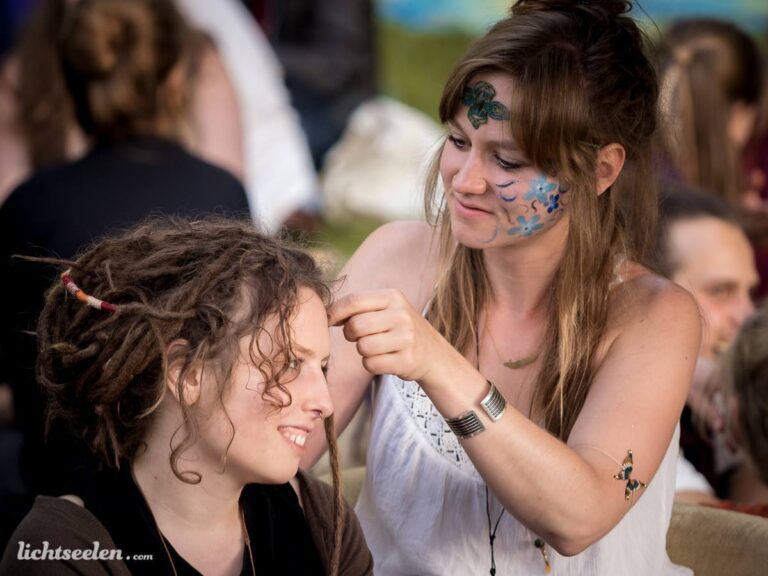 Zwei Frauen bei einem Festival, eine flechtet der anderen Zöpfe, umgeben von lebhafter, entspannter Atmosphäre.
