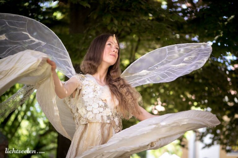 Eine elegante Frau mit großen, transparenten Flügeln posiert anmutig in einem grünen Garten.