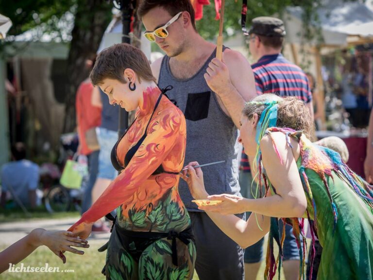 Frau mit farbenfrohen Bodypainting wird auf einem Festival von einer Künstlerin bemalt, im Hintergrund Menschen und Stände.