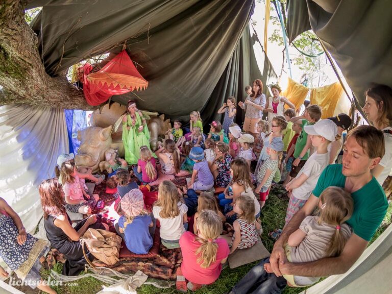 Eine fröhliche Gruppe von Kindern sitzt um eine Geschichtenerzählerin in einem zeltartigen Raum beim Festival.