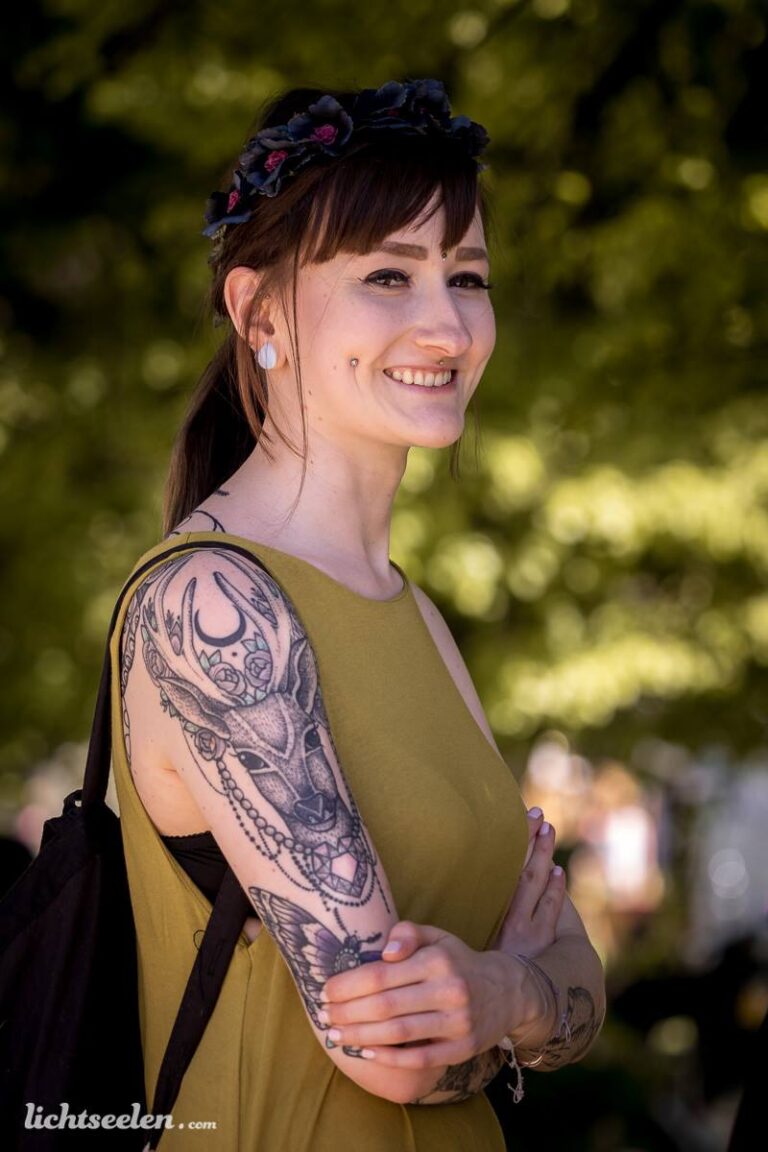 Lächelnde Frau mit Tattoos und Blumenkranz, aufgenommen vor grünen Bäumen, strahlt Freude aus.