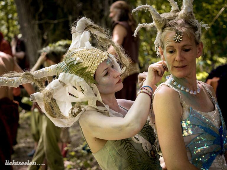 Eine Frau mit einem kunstvollen Kopfschmuck stylt eine andere Frau in einem festivalartigen Naturambiente.