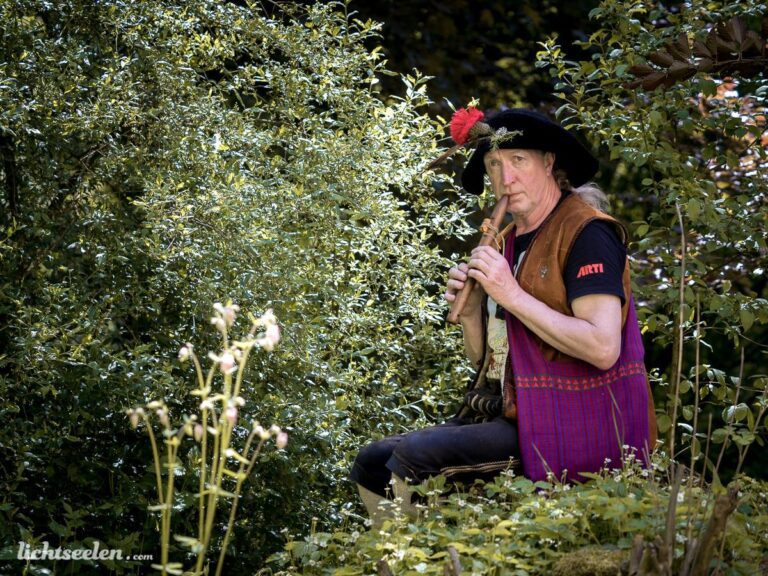 Musiker in traditioneller Kleidung spielt begeistert eine Flöte inmitten grüner Pflanzenlandschaft.