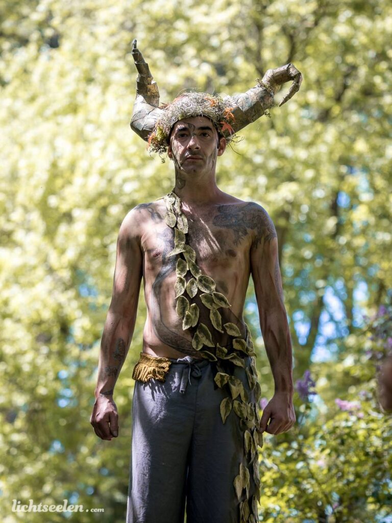 Eine personifizierte Naturfigur mit Geweih und Pflanzenkranz steht majestätisch im grünen Freien.