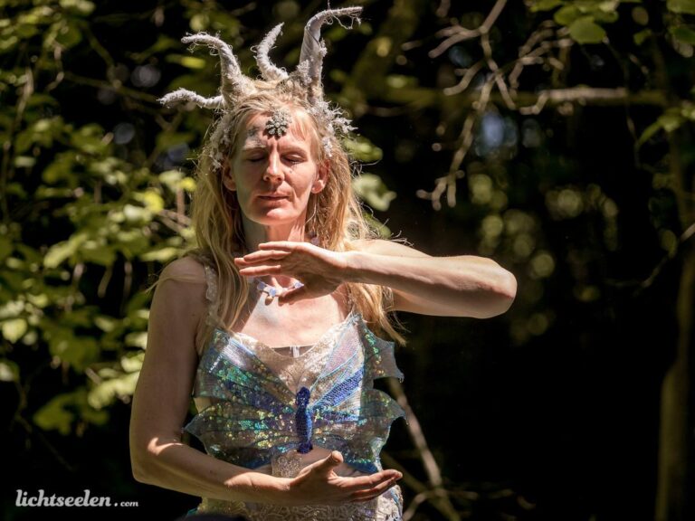 Eine Frau in fantasievoller Kleidung vollzieht eine anmutige Bewegung im Wald, umgeben von grüner Natur.