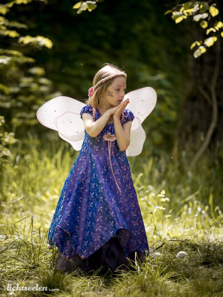 Ein Mädchen in einem lila Kleid mit Flügeln steht nachdenklich im Grünen, umgeben von Natur und sanften Licht.