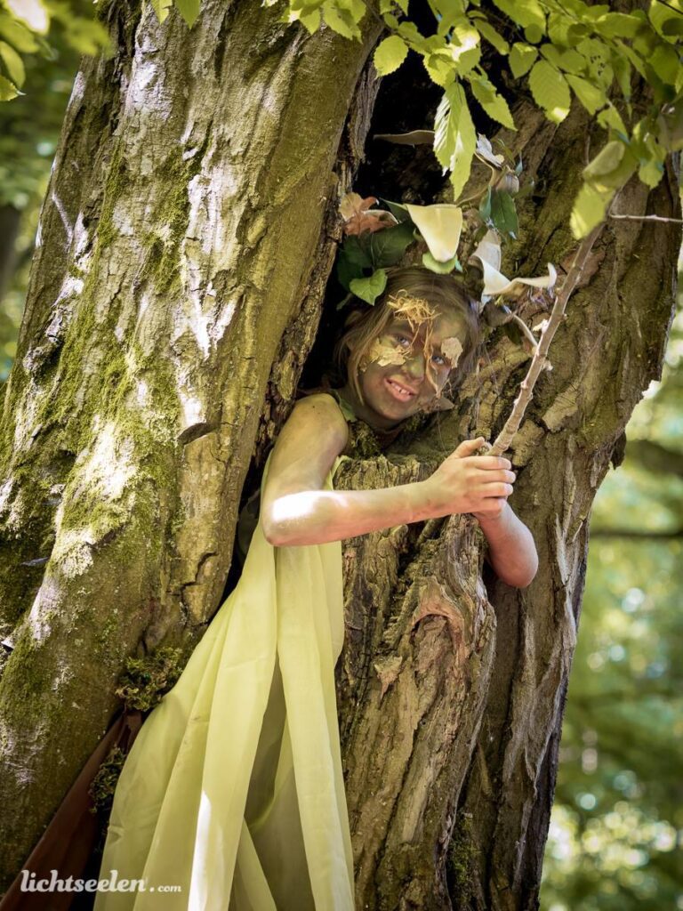 Ein Mädchen, verkleidet als Baumgeist, lächelt aus einem Baumstamm im sonnendurchfluteten Wald.