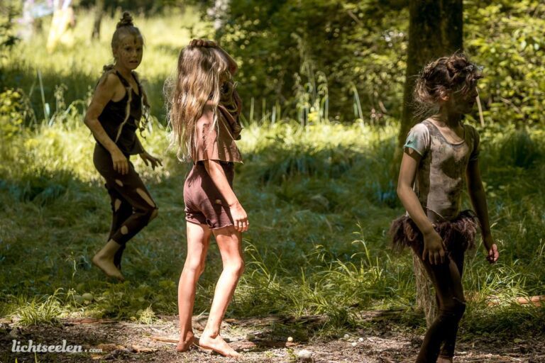 Drei spielende Kinder in natürlichen, erdigen Kostümen mit einem Waldhintergrund, die ausgelassen in der Sonne stehen.