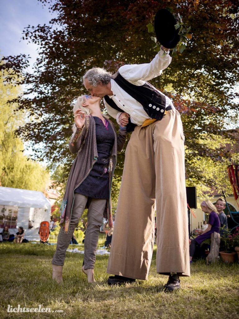 Ein attraktives Festival-Paar tanzt fröhlich, während ein großer Stelzenläufer ihnen Gesellschaft leistet.