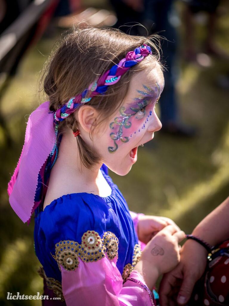 Mädchen mit buntem Kostüm und Gesichtsbemalung, strahlend und begeistert auf einem Festival.