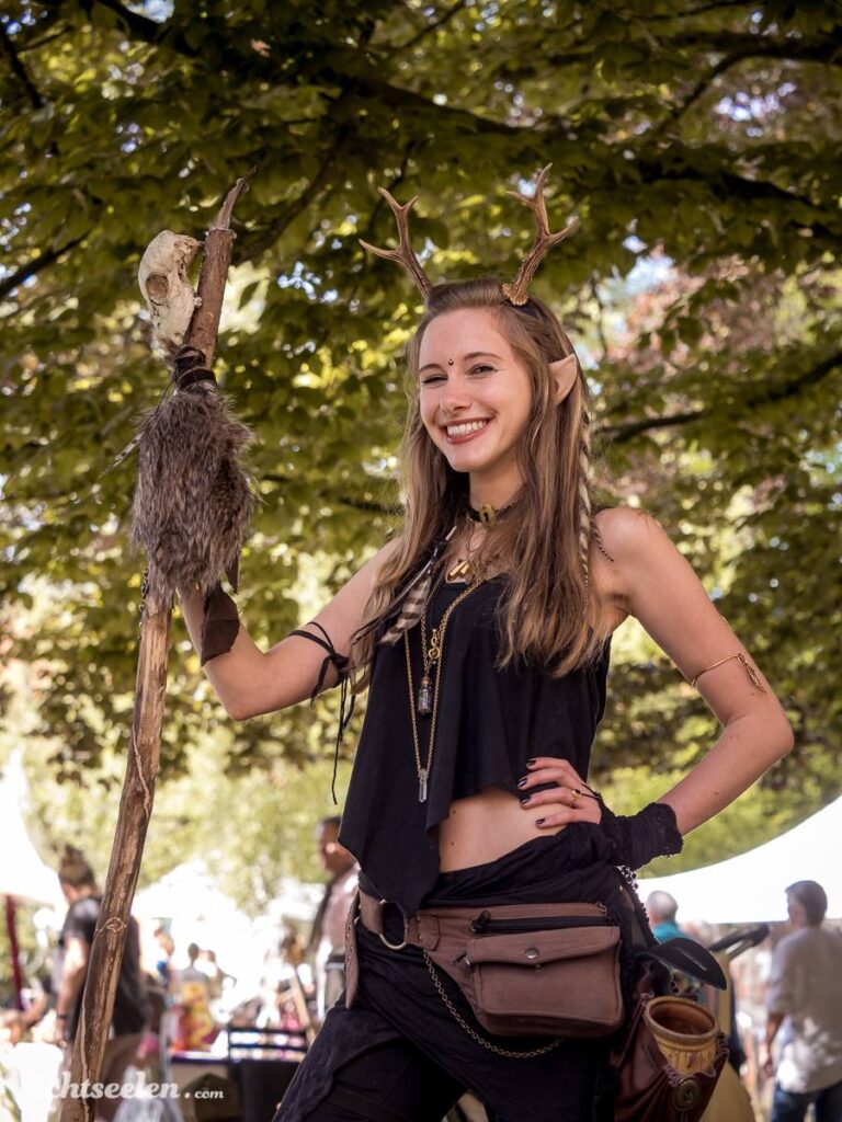 Frau im Fantasy-Kostüm mit Hörnern und einem Stock lächelt glücklich auf einem Festival unter Bäumen.