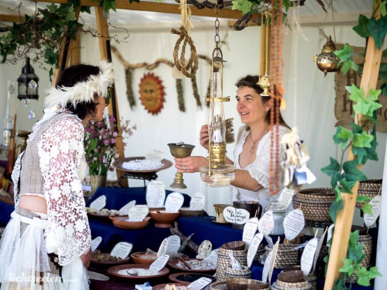 Zwei Frauen in festlicher Kleidung tauschen an einem Marktstand mit Naturprodukten in einem freundlichen, kreativen Ambiente.