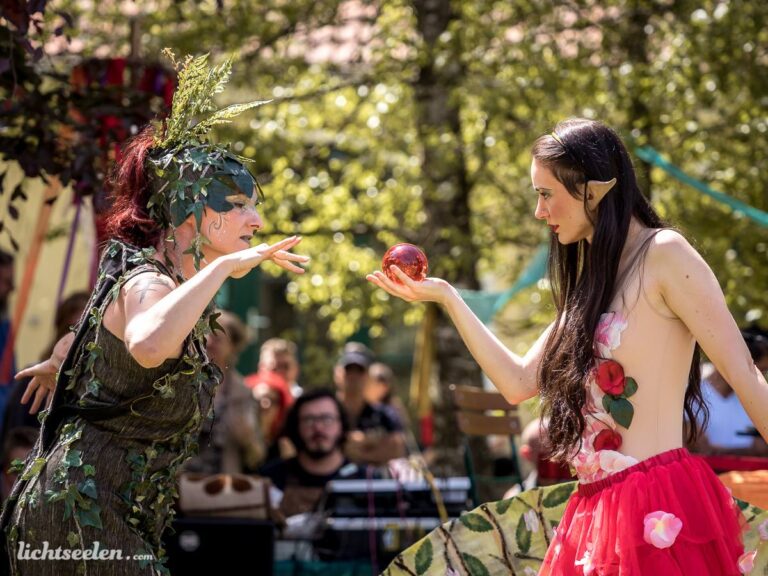 Zwei Darstellerinnen in fantasievoller Kleidung interagieren auf einem Festival, umgeben von Natur und fröhlichem Publikum.