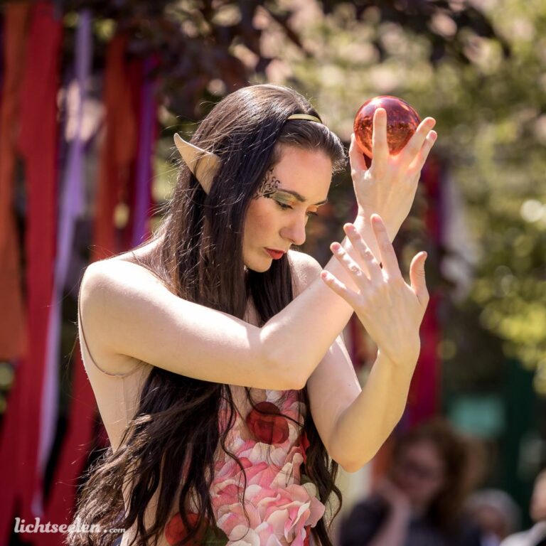 Eine elegante Frau mit Elfenohren vollzieht anmutige Bewegungen mit einer Glaskugel inmitten eines Festivalambientes.