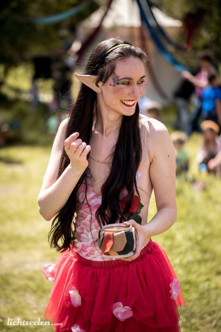 Elfenartige Frau mit langen Haaren und fröhlichem Lächeln, steht auf einer Wiese während eines Festivals.