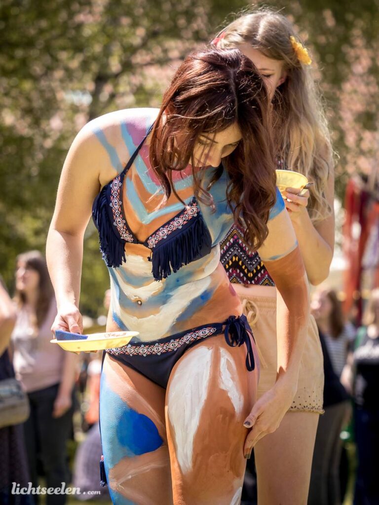 Zwei Frauen mit Körpermalerei, eine trägt Farbpalette, im Freien während eines Festivals.