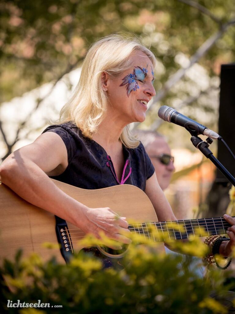 Frau mit Gesichtsbemalung spielt Gitarre und singt bei einem Festival im Freien.