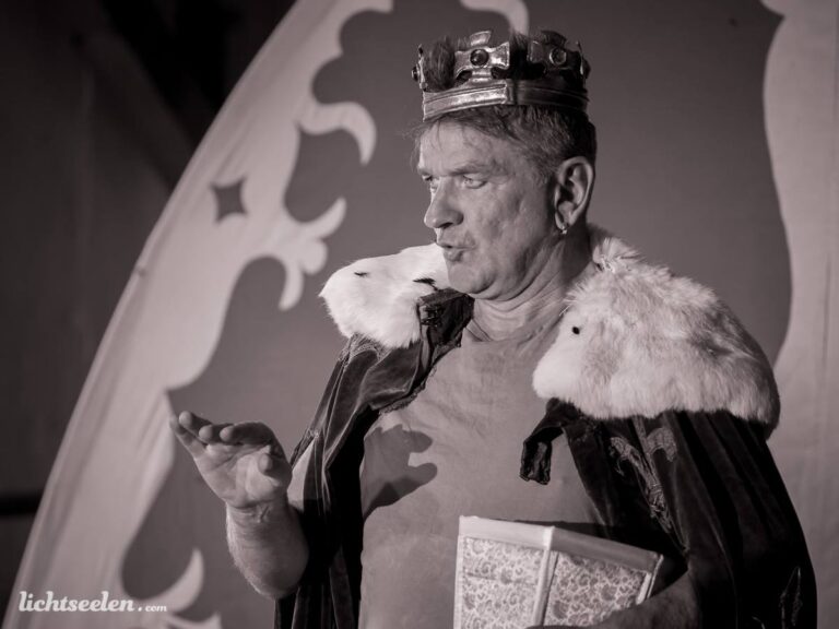 Ein Schauspieler in königlicher Robe mit Krone zeigt eine ausdrucksvolle Geste auf einer Bühne.