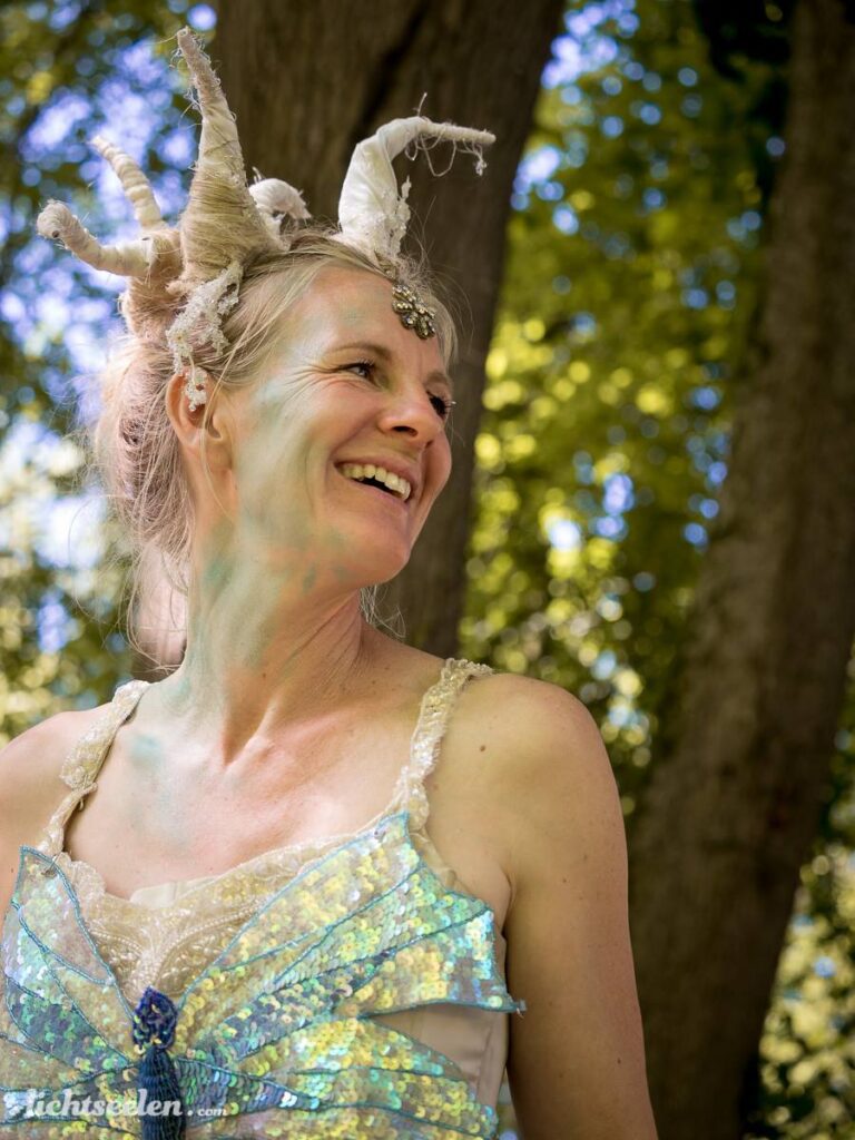 Lächelnde Frau mit Hornfrisur und glitzerndem Outfit vor grüner Naturkulisse, strahlt Freude und Magie aus.