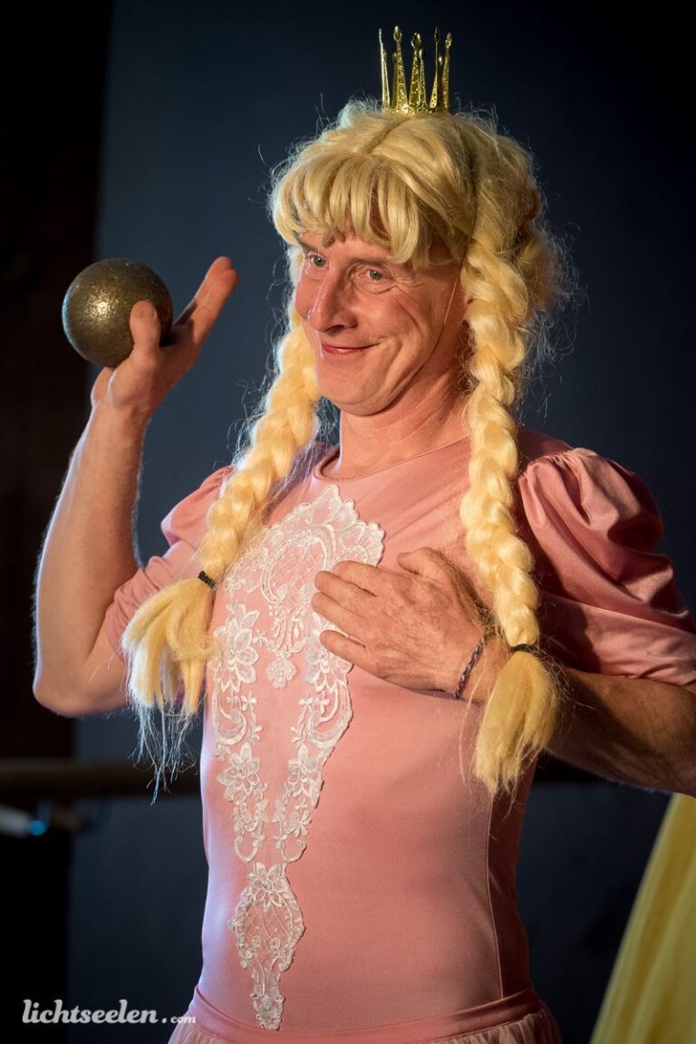 Ein Mann in einem rosa Kleid und mit blonden Zöpfen hält glänzende Kugel und strahlt Freude aus.