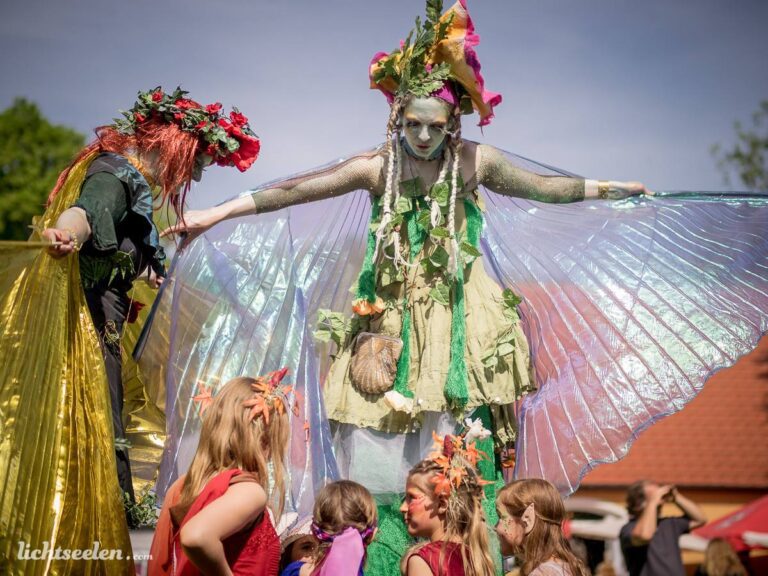 Fantasievoller Auftritt einer kunstvoll geschminkten Darstellerin mit Flügeln und bunten Kindern auf einem Festival.
