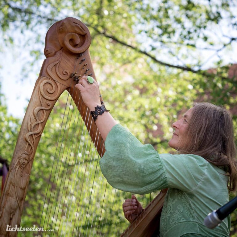 Frau in grüner Bluse spielt an ihrer kunstvoll verzierten Harfe unter Bäumen bei einem Festival.