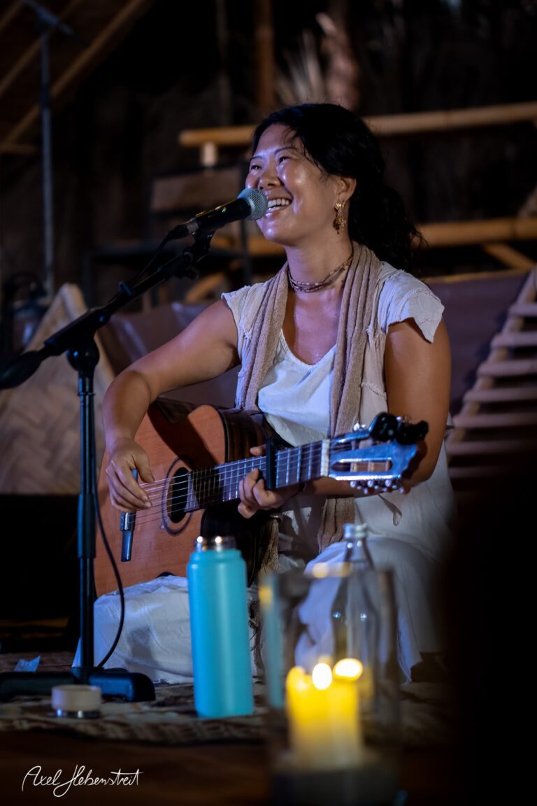 Eine lächelnde Musikerin spielt mit einer Gitarre in einer stimmungsvollen Atmosphäre während eines Konzerts – © Axel Hebenstreit.