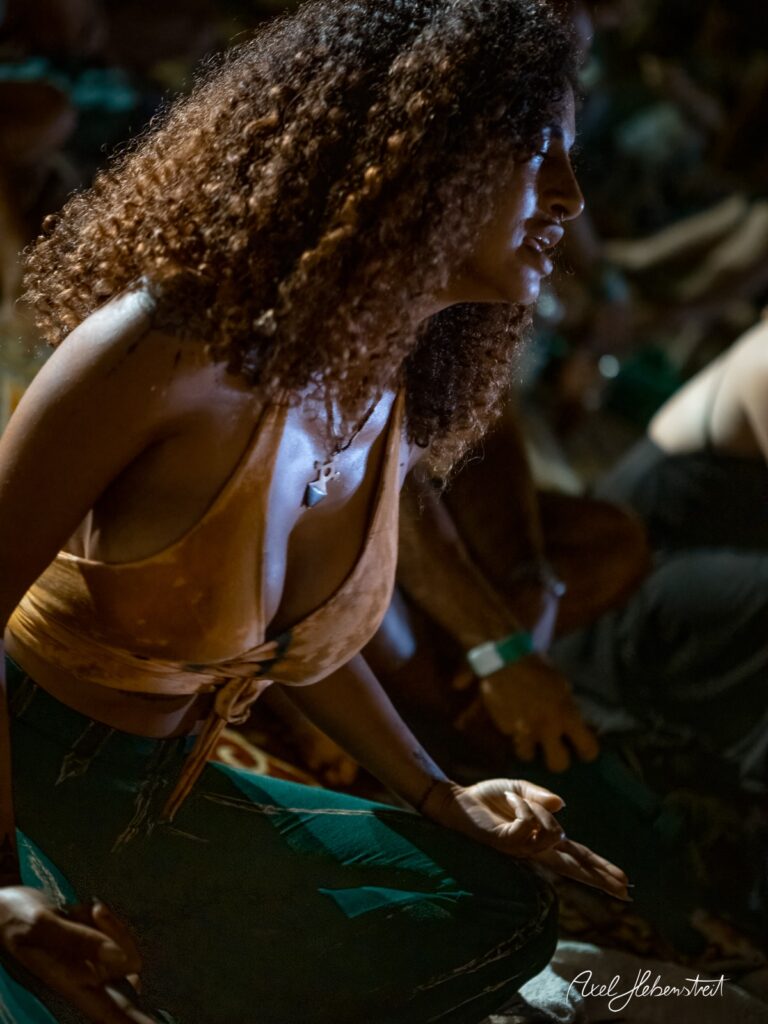 Eine junge Frau mit lockigem Haar in meditativer Pose auf einem Festival im Dämmerlicht – © Axel Hebenstreit