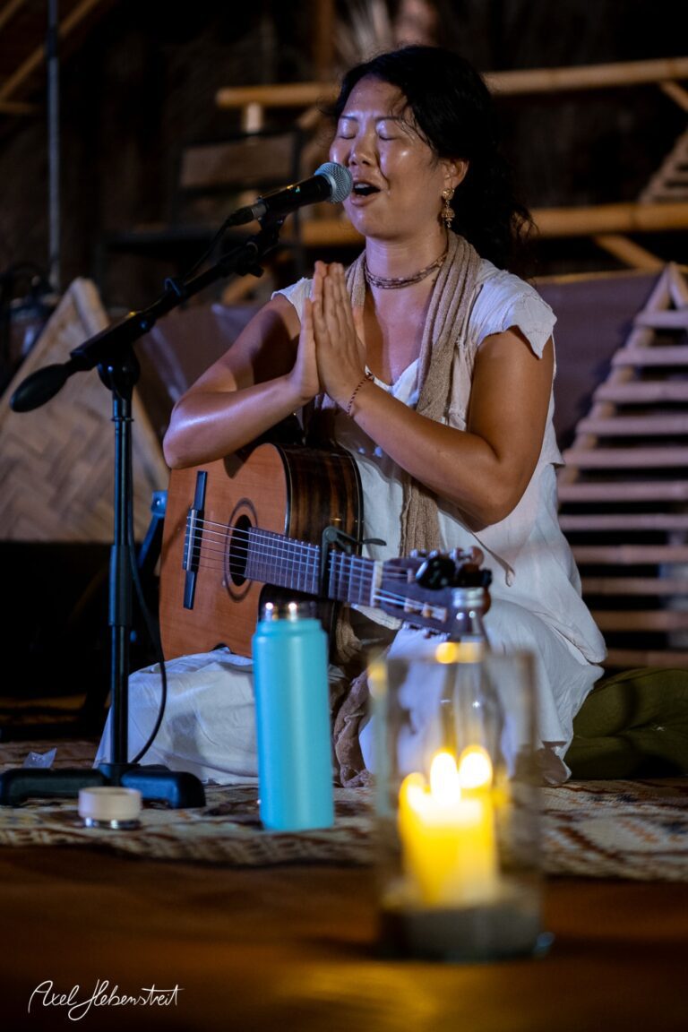 Eine Musikerin in weißem Kleid singt und spielt Gitarre, während sie andächtig mit geschlossenen Augen im Licht einer Kerze sitzt – © Axel Hebenstreit