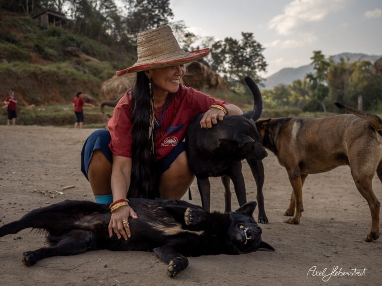 Eine lächelnde Frau in traditioneller Kleidung streichelt einen Hund auf einem staubigen Platz in der Natur. – © Axel Hebenstreit